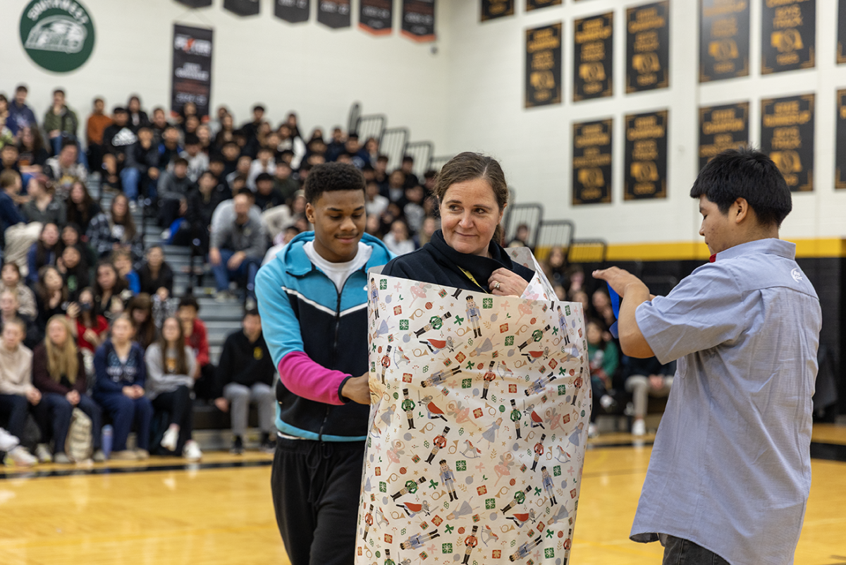 Malcolm Wilson (left) and Gasparin Ramos Larios (right) wrap English teacher Stacey Smith in Christmas wrapping paper during a game at the winter pep rally in the Al Bahe Gym.